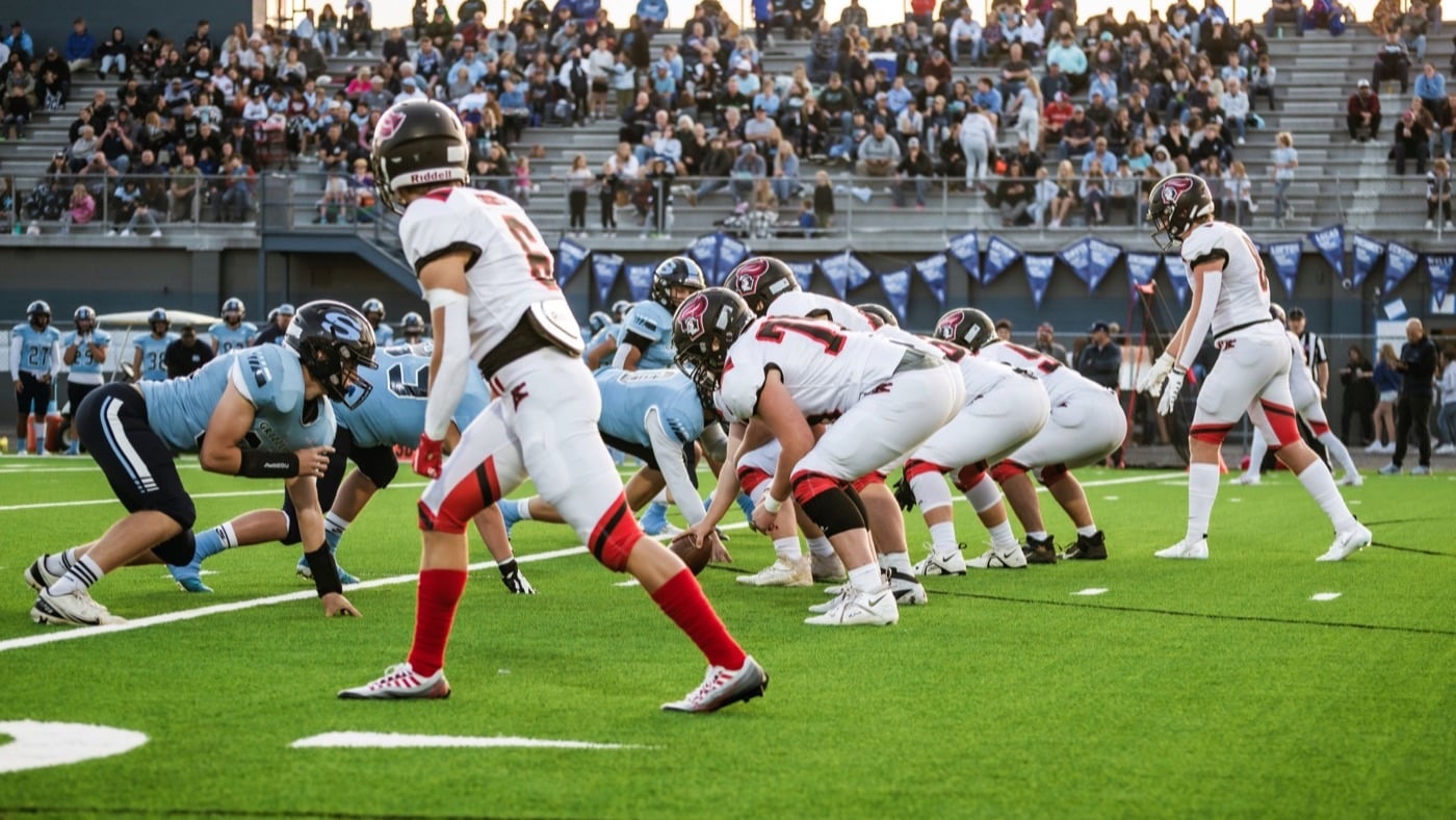 Two high school football teams lined up at the line of scrimmage with spectators in the stands