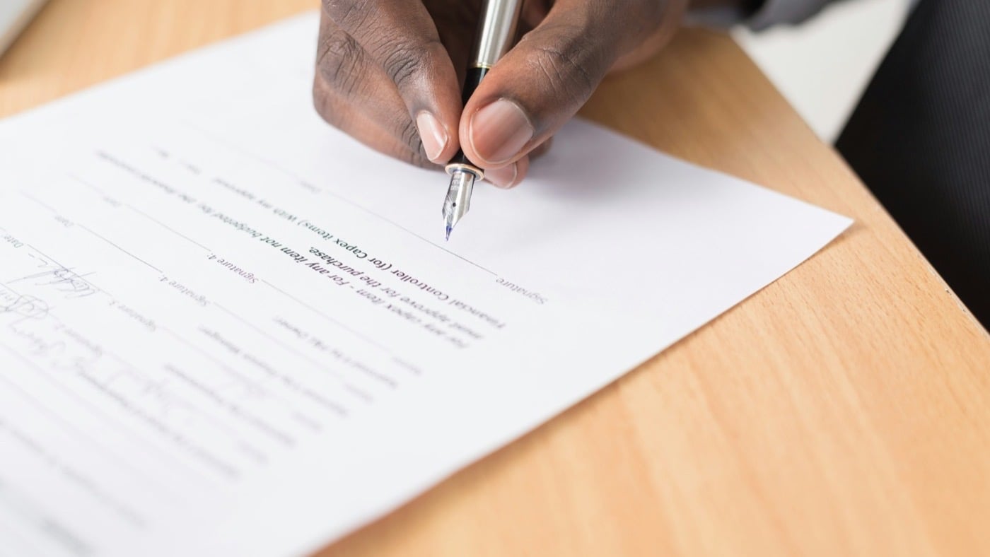A hand signing a printed financial approval form on a wooden desk