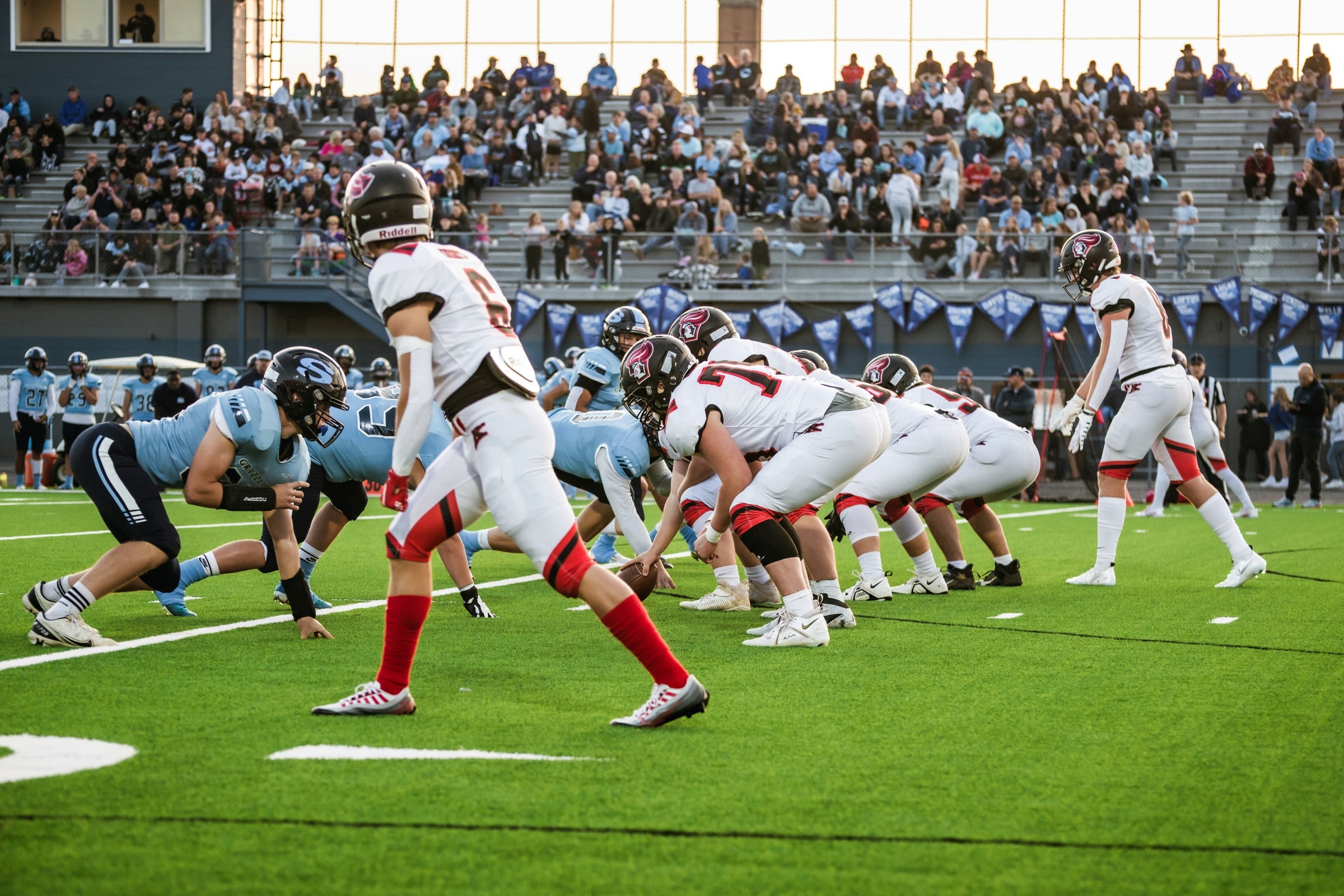 Two high school football teams lined up at the line of scrimmage under stadium lights, with a full crowd in the stands behind them